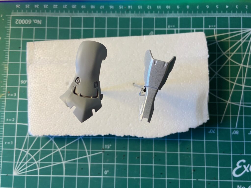 Two gundam parts side by side, one being a flat grey colour and the other a shiny silver colour.